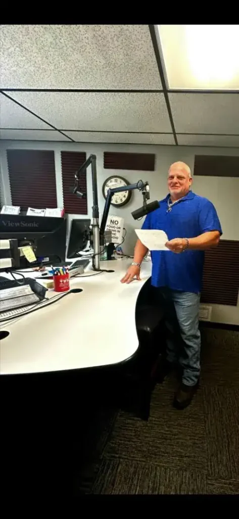 A man in a blue shirt stands at a radio station desk holding papers, with microphones, computer monitors, and a wall clock visible in the background, ready to discuss pest control Hernando & Pasco County, FL.